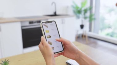 Kafkasyalı bir kadın modern mutfakta online yemek siparişi vermek için akıllı telefon kullanıyor. Sandviçler ve meyve suları gibi çeşitli yemek seçenekleri içeren dağıtım uygulaması menüsü.
