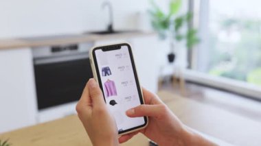 Beyaz bir kadın akıllı telefon kullanarak internetten kıyafet alışverişi yapıyor. Görüntü, teknolojiye odaklı online alışverişin kolaylığını betimliyor. Telefon tutan eller kıyafet seçenekleriyle birlikte web sayfasını göster.