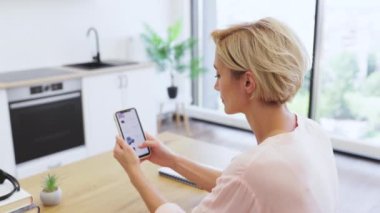 30 'lu yaşlarda beyaz bir kadın akıllı telefon kullanarak online kıyafet alışverişi yapıyor. Parlak, modern mutfakta. Tahta masada oturuyor, internet sitesinde geziniyor..