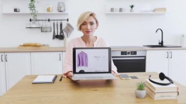 Gülümseyen beyaz bir kadın online kıyafet alışveriş sitesiyle dizüstü bilgisayar sergiliyor. Modern mutfak ortamında 30 'lu yaşların ortalarında sarışın bir kadın internet moda alışverişi yapıyor..
