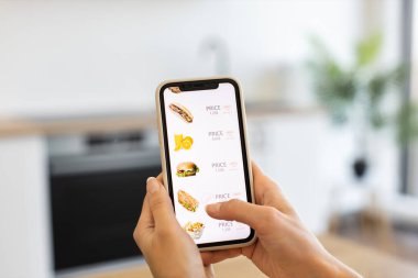Kafkasyalı bir kadın modern mutfakta online yemek siparişi vermek için akıllı telefon kullanıyor. Sandviçler ve meyve suları gibi çeşitli yemek seçenekleri içeren dağıtım uygulaması menüsü.
