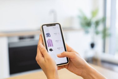 Beyaz bir kadın akıllı telefon kullanarak internetten kıyafet alışverişi yapıyor. Görüntü, teknolojiye odaklı online alışverişin kolaylığını betimliyor. Telefon tutan eller kıyafet seçenekleriyle birlikte web sayfasını göster.