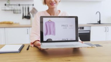 Elinde dizüstü bilgisayarla gülümseyen beyaz bir kadın online alışveriş sitesinde kıyafetlerini gösteriyor. Dizüstü bilgisayarda, fiyat ve indirim teklifiyle birlikte pembe sweatshirt gösteriliyor.