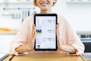 İnternette kozmetik dağıtım sitesi gösteren tablet tutan mutlu beyaz kadın. Tablet ekranda indirimli ve indirimli çeşitli cilt ürünleri gösteriliyor, uygun alışveriş deneyimleri vurgulanıyor.