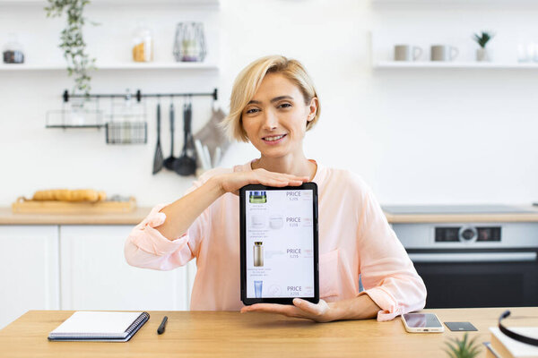 Happy Caucasian woman holding tablet displaying online cosmetics delivery website in modern kitchen. Middle-aged female demonstrates digital shopping experience, emphasizing convenience and technology
