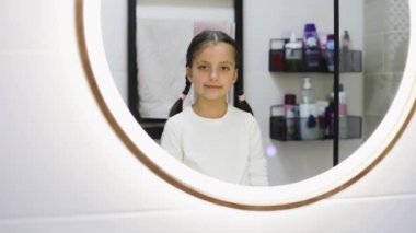 Saçları örgülü ve dişleri dökülmüş beyaz bir kız banyoda dikiliyor. Kameraya gülümsüyor, cazibesini ve masumiyetini sunuyor. Çocukluk, büyüme ve aile anları temaları için ideal.