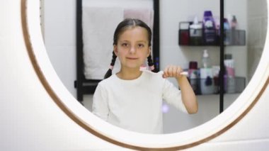 Saçları örgülü, dişlerini fırçalayan ve banyo ortamında aynaya gülümseyen beyaz bir kız. Eksik süt dişlerini göstermek ve günlük diş hijyeni rutinine başlamak.