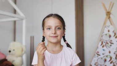 Saçı örgülü mutlu, beyaz bir kız. Ev ortamında bebek dişlerini gösteriyor. Heyecanını dile getiriyor..