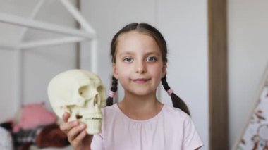 Kafkasyalı bir kız, bebek dişleri kayıp ve kafatası modelini içeride tutuyor. Ev ortamında merakı ve oyunculuğu ifade eder. Saç stili at kuyruğu eğitici temaya cazibe katar.
