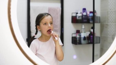 Banyo aynasına bakarken dişlerini fırçalayan beyaz bir kızın portresi, iyi ağız temizliği alışkanlıklarını vurguluyor..