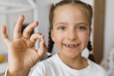 Çocukluk dönüm noktalarını sembolize eden, düşmüş bebek dişi sergileyen heyecanlı beyaz bir kız. Gülümseyen çocuk diş kaybetmekten zevk alıyor, ev ortamını ve büyümeyi vurguluyor.