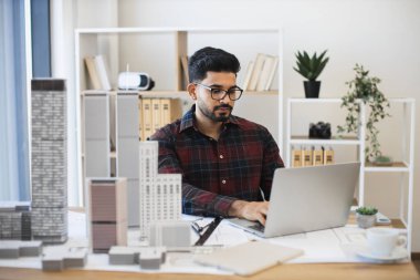 Genç Hintli erkek mimar dizüstü bilgisayarda çalışıyor, masa başında modeller inşa ederek modern şehir planı tasarlıyor..