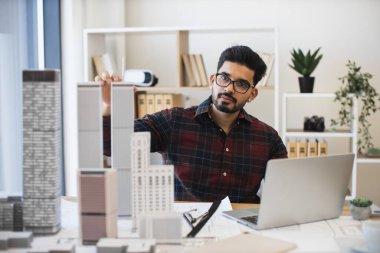 Genç Hintli erkek mimar dizüstü bilgisayar ve ölçekli modellerle şehir planlama üzerinde çalışıyor. Modern ofis ortamında profesyonelce çalışmaya odaklanmış. Mimarlık, yaratıcılık, kentsel planlama kavramı.