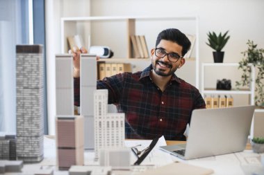 Genç Hintli erkek mimar dizüstü bilgisayar ve ölçekli modellerle şehir planlama üzerinde çalışıyor. Modern ofis ortamında profesyonelce çalışmaya odaklanmış. Mimarlık, yaratıcılık, kentsel planlama kavramı.