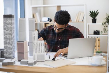 Hintli erkek mimar kalem ve kağıtla modern şehir planını çiziyor. Stüdyoda modeller ve dizüstü bilgisayarlarla mimari proje üzerinde çalışan genç bir profesyonel..