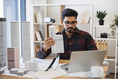 Genç Hintli adam ofis ortamında mimari model tasarlıyor. Erkek mimar, yenilikçi kentsel planlama için dizüstü bilgisayar ve fiziksel yapı modellerini kullanarak modern şehir modeline odaklanır..