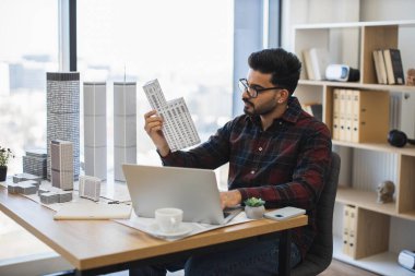 Genç Hintli erkek mimar modern şehrin modelini yapıyor. Bina modelleri ve ofis ortamında dizüstü bilgisayarlarla çalışıyor..