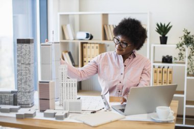 Pembe gömlekli Afrikalı kadın mimar modern ofis ortamında şehir manzarası modeli tasarlıyor, yaratıcılık ve hassasiyet sergiliyor. Düşünceli ifadesi sanatına olan bağlılığını vurguluyor..