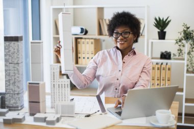 Afrikalı kadın mimar modern şehir modelini iş başında tasarlıyor. Gözlüklü genç profesyonel çalışma masasında planlar, dizüstü bilgisayarlar, mimari maketlerle çalışıyor. Parlak ofis ayarları, yaratıcılığa odaklan.