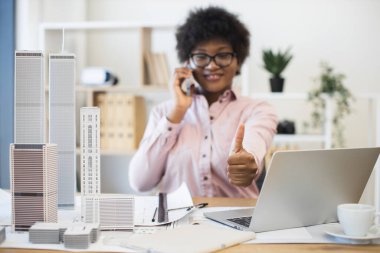 Kendine güvenen Afrikalı iş kadını mimar şehir projesiyle ilgili telefon görüşmesi yapıyor. Ofiste dizüstü bilgisayar ve mimari modellerle çalışırken gülümseyen ve başparmak kaldıran.