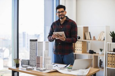 Hintli erkek mimar, yaratıcı çalışma alanında dijital tablet kullanarak modern şehir projesini geliştirir. Masadaki uzun bina modelleri yenilikçi şehir tasarımını sembolize ediyor. Yaş grubundaki gençlerden orta yaşlılara.