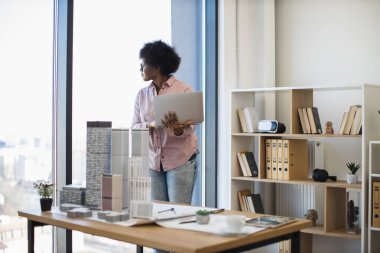 Afrikalı Amerikalı kadın mimar dijital dizüstü bilgisayar kullanarak modern şehir için proje geliştirir. Masanın üzerinde dizüstü bilgisayarla duruyor, yaratıcılığı ve yeniliği yansıtıyor..