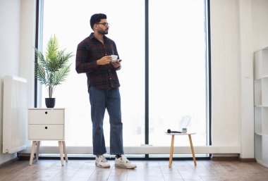 Elinde kahve fincanı olan Hintli bir erkek minimalist oda ortamında pencereden dışarı bakıyor. İç mekan ve ahşap mobilyalarla rahatlama ve modern yaşam tarzını ön plana çıkarıyor.