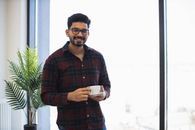 Parlak ofis ortamında elinde kahve tutarken gülümseyen günlük kıyafetli genç Hintli adam. Kapalı alan ve geniş pencere rahat ve dostane bir atmosfer yaratıyor. Gözlükler modern tarzın dokunuşunu ekler.