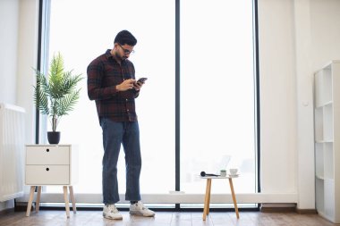 Genç Hintli adam parlak ofis ortamında akıllı telefon kullanıyor. Doğal ışık, küçük masa ve kapalı bitkiler ile modern minimalist dekor barışçıl çalışma ortamı oluşturur.