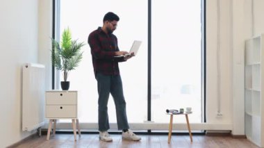 Genç Hintli erkek, modern ofiste dizüstü bilgisayarla pencerenin önünde duruyor. Flanel gömlek ve kotla günlük kıyafet. Parlak ve minimalist çalışma alanı. Kapalı bitki ve doğal ışık..