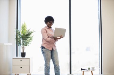 Gündelik kıyafet giymiş Afro-Amerikalı iş kadını dizüstü bilgisayarla çalışan pencere kenarında duruyor. Parlak modern ofis, kapalı alanda profesyonel ama rahat çalışma alanı oluşturur..