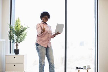 Sıradan bir ofiste gülümseyen Afrikalı Amerikalı kadın, elinde dizüstü bilgisayarla, baş parmak hareketleriyle, pencerenin yanında duruyordu. Saksı bitkisi ve küçük masalı parlak modern çalışma alanı.