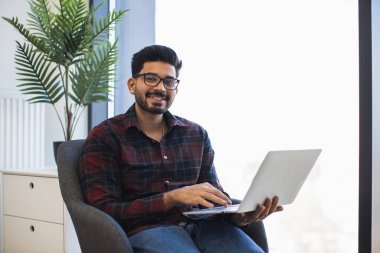 Elinde dizüstü bilgisayarla ofis koltuğunda oturan gülümseyen genç Hintli adam. Doğal ışık alan sıradan bir çalışma ortamı. Rahat ve rahat bir atmosfer. Yeşilliği olan modern çalışma alanı.