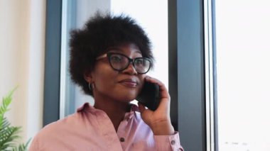 Ofis ortamında pencere kenarındaki telefon görüşmelerine katılan, günlük kıyafetli Afrikalı Amerikalı kadın, güven ve profesyonelliği iletiyor..