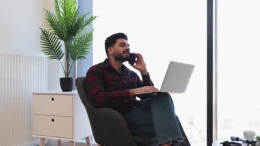 30 'lu yaşlarda Hintli bir adam evde bilgisayarla ve telefonla çalışıyor. Kapalı alan bitkisi ve doğal ışık olan rahat bir ortam. Uzak çalışma ve üretkenlik kavramı.