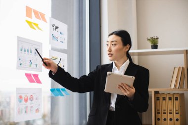 Modern ofis ortamında cam duvarda tablet ve kalem kullanan Asyalı iş kadını..