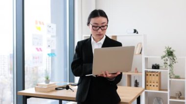 Modern ofis ortamında dizüstü bilgisayar tutan Asyalı kadın iş kadını. Odaklanmış ve meşgul görünüyor, büyük pencere manzaralı masa başında duruyor, kurumsal atmosferi gösteriyor..