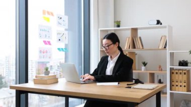 Modern parlak ofis ortamında dizüstü bilgisayarla çalışan profesyonel giyimli Asyalı bir iş kadını. Verimliliği ve profesyonelliği vurgulayan şık bir ortamda çalışmaya odaklanan genç bir kadın.