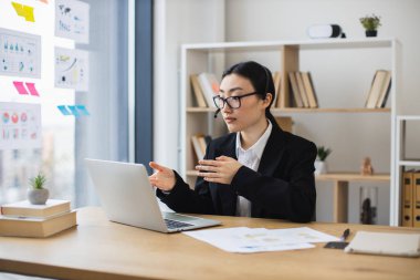 Siyah takım elbiseli Asyalı iş kadını video görüşmesi sırasında kulaklığı olan bir laptop kullanıyor. Genç profesyonel, kitap rafı ve bitki örtüsüyle modern ofis ortamında çevrimiçi toplantıya katılıyor..