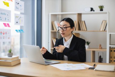 Siyah takım elbiseli Asyalı iş kadını video görüşmesi sırasında kulaklığı olan bir laptop kullanıyor. Genç profesyonel, kitap rafı ve bitki örtüsüyle modern ofis ortamında çevrimiçi toplantıya katılıyor..