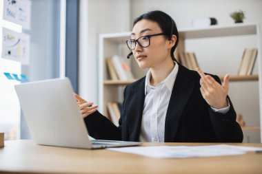 İnternetteki toplantıya katılan kulaklık takan Asyalı iş kadını. Modern ofis ortamında profesyonel kıyafet. İletişimi kolaylaştıran teknoloji.