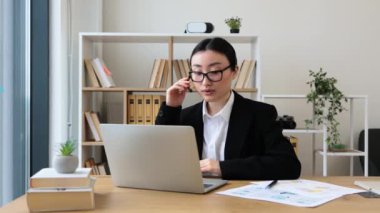 İnternetteki toplantıya katılan kulaklık takan Asyalı iş kadını. Modern ofis ortamında profesyonel kıyafet. İletişimi kolaylaştıran teknoloji.
