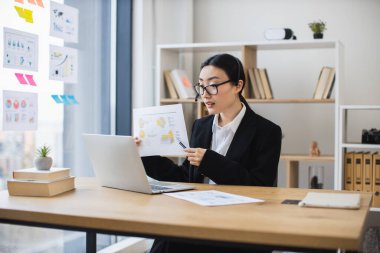 Resmi giyinmiş Asyalı kadın profesyonel, online toplantı düzenliyor. Analiz raporunu dizüstü bilgisayardan sunuyor. Modern ofis ayarları üretkenlik, iletişim, uzmanlık gösteriyor.