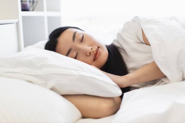 Asyalı bir kadın rahat bir yatakta uyuyor, beyaz çarşaflarla çevrili, huzuru ve huzuru yakalıyor. Rahatlamanın ve huzurlu bir atmosferin mükemmel tasviri..