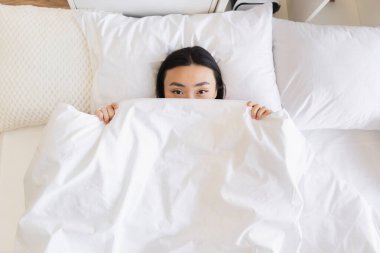 Beyaz battaniyenin altında saklanan Asyalı bir kadın. Rahat ve rahat görünüyor. Parlak, havadar odada sıcaklık ve sükunet hissi yaratıyor..