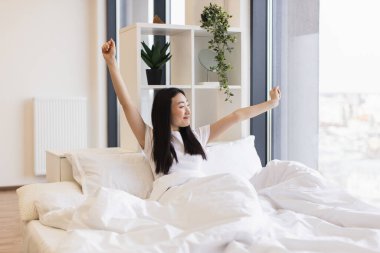 Asyalı bir kadın aydınlık yatak odasında uyanıyor, gülümsüyor ve kollarını açıyor, sabahtan zevk alıyor. Yatak çarşafları beyaz, oda modern dekora sahip. Rahatlama ve pozitiflik yaşayan genç bir kadın..