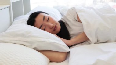 Asyalı bir kadın rahat bir yatakta uyuyor, beyaz çarşaflarla çevrili, huzuru ve huzuru yakalıyor. Rahatlamanın ve huzurlu bir atmosferin mükemmel tasviri..