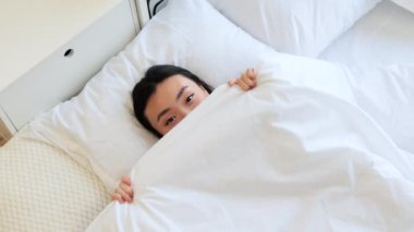 Beyaz battaniyenin altında saklanan Asyalı bir kadın. Rahat ve rahat görünüyor. Parlak, havadar odada sıcaklık ve sükunet hissi yaratıyor..