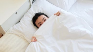 Yatakta gülümseyen Asyalı kadın mutluluk ve rahatlama gösteriyor. Genç bir kadınla kapalı bir sabah sahnesi, parlak bir yatak ve neşeli bir ifade, Asyalı bir kadın yeni günü neşe ve olumlulukla karşılıyor..