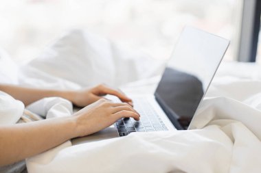 Genç Asyalı kadın rahat ev ortamında uzaktan çalışmak için dizüstü bilgisayar kullanıyor. Rahat duruş rahatlık ve odaklanmayı önerir. Eller klavyede üretkenliği ve modern yaşam tarzını yansıtır.